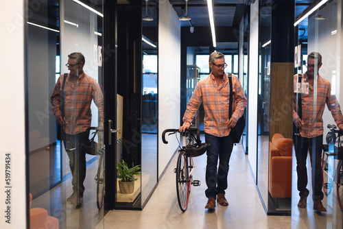 Fotografia, Obraz Caucasian mature businessman walking with bicycle in corridor of creative office