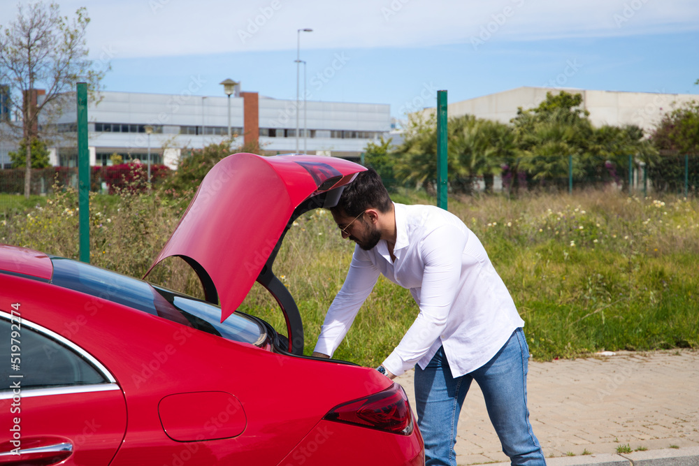 Young, handsome man opens the boot of his red sports car. The man is wealthy. High standard of living and well positioned financially. Young, handsome man opens the boot of his red sports car. The man is wealthy. High standard of living and well positioned financially.