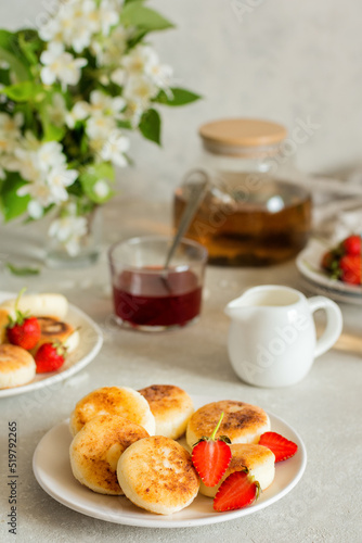 Cheesecakes with strawberries on a white plate on a table with flowers and tea. Healthy breakfast on a sunny day.