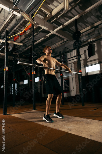 Portrait of shirtless sportive young man training, lifting heavy barbell, doing exercises isolated over gym background