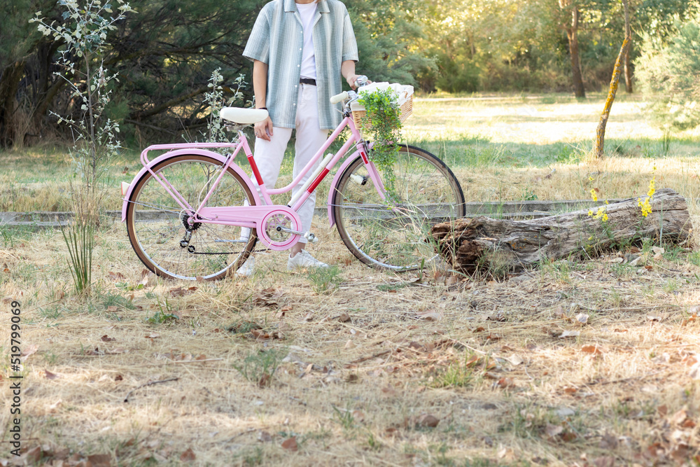 Fototapeta premium Cropped side view of unrecognizable young fashinable man holding a pink bicycle with a wicker basket and plants