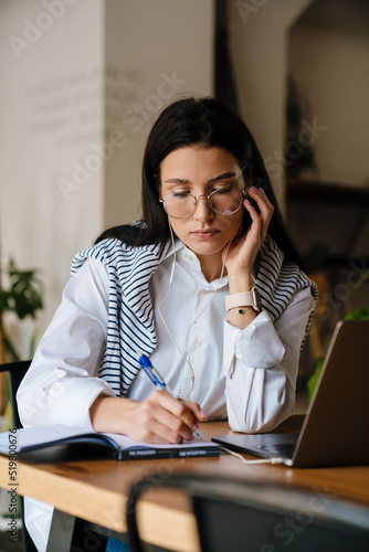 White woman writing down notes while working with laptop at cafe