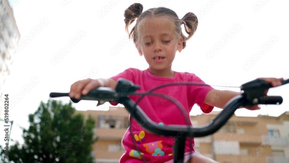 Cute little smiling girl riding bike bicycle in city on parking sunny summer day. Active family leisure with kids. Little caucasian girl 5 years old have fun with pink child bicycle Cute little smiling girl riding bike bicycle in city on parking sunny summer day. Active family leisure with kids. Little caucasian girl 5 years old have fun with pink child bicycle