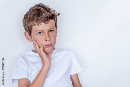 Upset white Eastern European 9 year old boy in a white t-shirt. The child looks sadly directly into the camera, involuntarily raising his hand to his face.