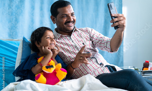 Fotografia Happy smiling father with recovered sick daughter making video call on mobile phone at hospital ward - concept of technology, relationship and health care