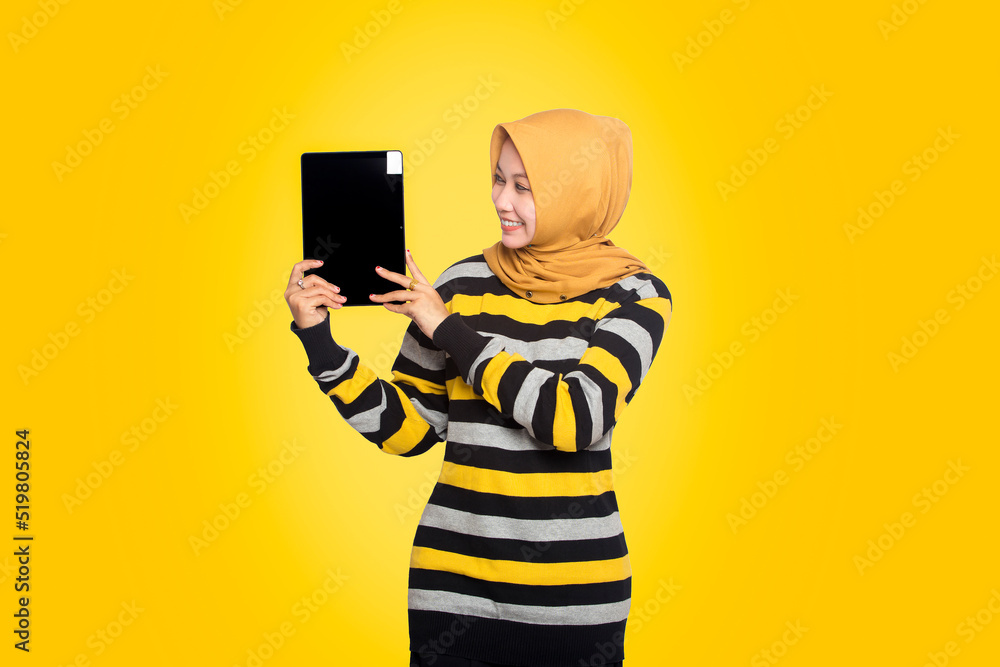 Happy millennial Asian beautiful woman showing off digital computer tablet. Beautiful Muslim teenage girl wearing hijab happy in striped dress using tablet computer, isolated on yellow background. Happy millennial Asian beautiful woman showing off digital computer tablet. Beautiful Muslim teenage girl wearing hijab happy in striped dress using tablet computer, isolated on yellow background.
