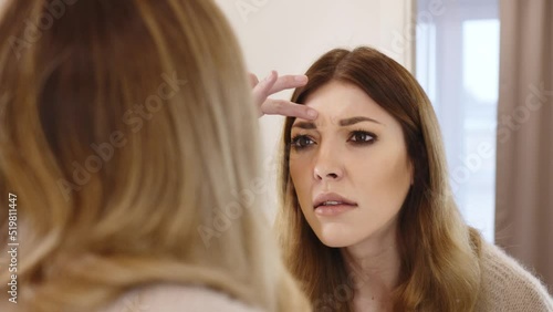 A young woman worried about the condition of her skin. He is looking at the new wrinkles on his face in the bathroom mirror.