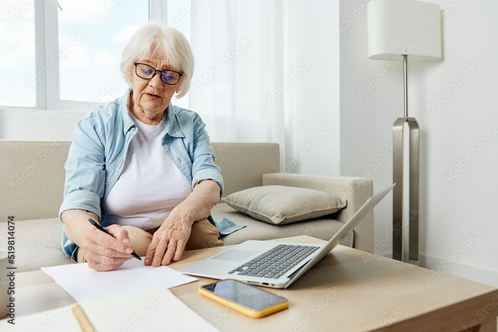A mature elderly woman in a bright apartment is sitting at a table on the sofa using a laptop. Active retirement lifestyle. The concept of studying leisure. A mature elderly woman in a bright apartment is sitting at a table on the sofa using a laptop. Active retirement lifestyle. The concept of studying leisure.