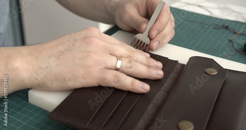 Mens hand holding a hammer and hole punch and makes a holes for seawing a leather wallet in his workshop. Working process with a brown natural leather. Craftsman holding a crafting tools.
