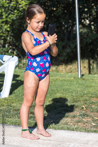 Little girl in a swimsuit, standing on the edge of the pool, looking at the mobile phone. Childhood, children, smartphone, technology, vacation, internet and fun concept.