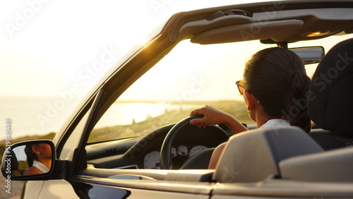 Woman alone in a black convertible car looks at the sunset in front of the sea and is moved by how wonderful it is, she feels free and happy during her vacation alone, independent and strong woman