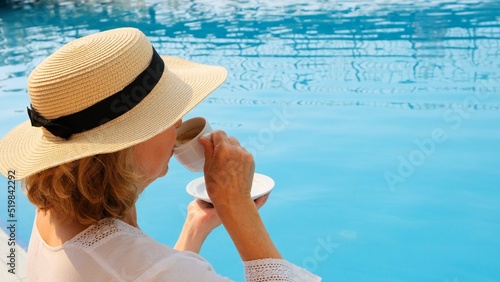 Close up of a woman aged 50-55 in a straw hat drinking from a cup of coffee next to a blue swimming pool, luxurious good morning, start of the day