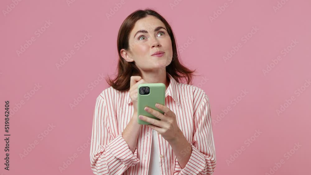 Young dreamful pensive minded woman 20s she wearing shirt t-shirt hold using mobile cell phone typing browsing chatting send sms isolated on plain pastel light pink background People lifestyle concept Young dreamful pensive minded woman 20s she wearing shirt t-shirt hold using mobile cell phone typing browsing chatting send sms isolated on plain pastel light pink background People lifestyle concept