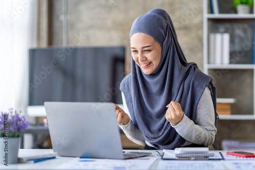 Happy Muslim businesswoman wearing hijab Muslim female office employee looking at laptop screen, celebrating success