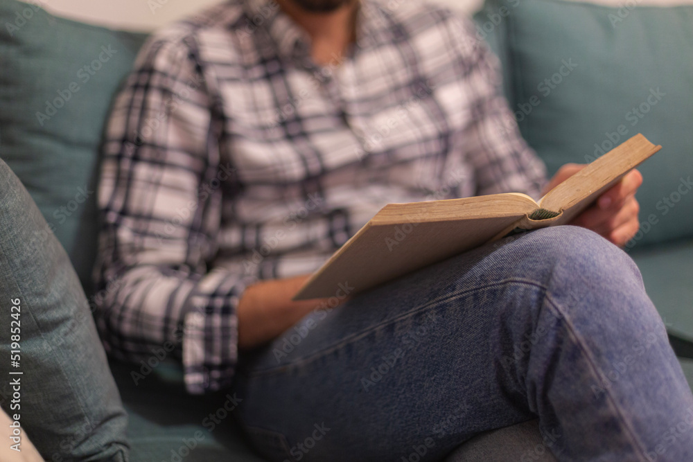 Fototapeta premium Closeup view of male hands holding book. Young man relaxing at home sitting in a couch. Selective focus on book, blurred background. Horizontal.