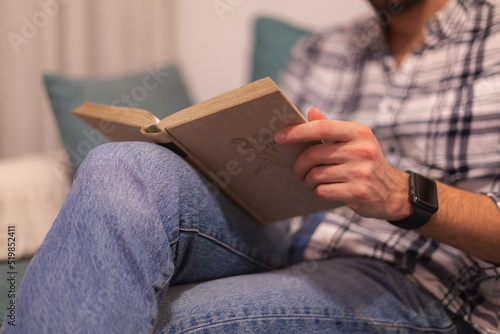 Closeup view of male hands holding book. Young man relaxing at home sitting in a couch. Selective focus on hand, blurred background. Horizontal.