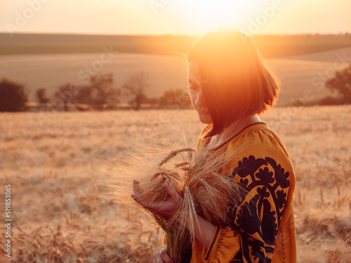 portrait of young woman in Ukrainian traditional clothes walking across golden field holding heap of rye by sunset light, copy space