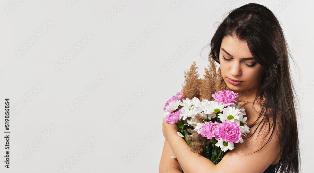Amazing brunette serious lady keeping bouquet of flowers to hide her breast naked and looking at the flowers.