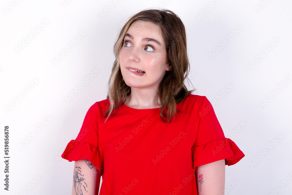 Amazed young caucasian woman wearing red T-shirt over white background bitting lip and looking tricky to empty space. Amazed young caucasian woman wearing red T-shirt over white background bitting lip and looking tricky to empty space.