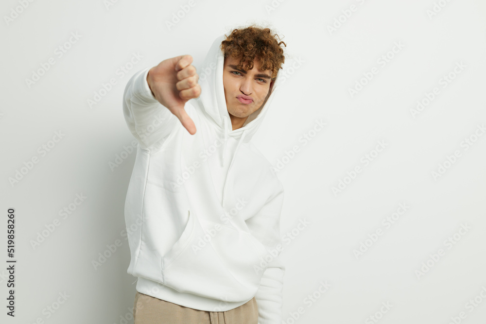 a cute, attractive man stands on a white background in a white hoodie and gives a thumbs down. Horizontal photo with an empty space for inserting an advertising layout