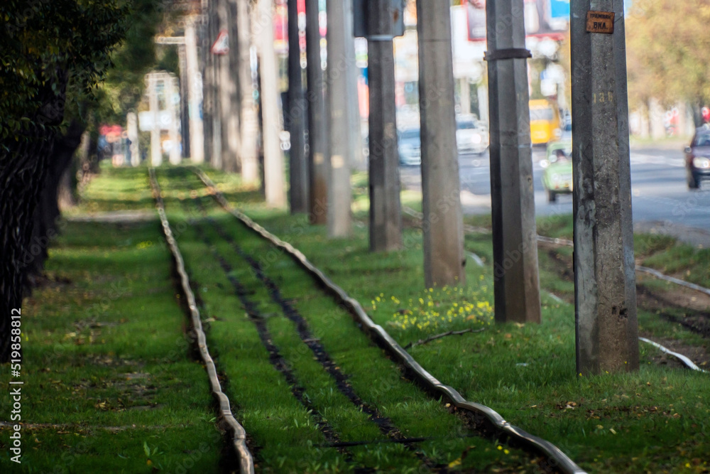 Crooked tram rails among green grass, trees and electric lampposts against the background of an asphalt road Crooked tram rails among green grass, trees and electric lampposts against the background of an asphalt road