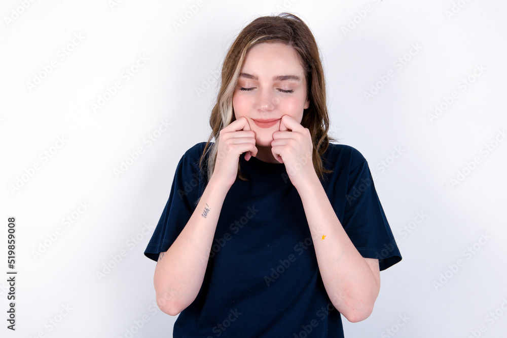 Pleased young caucasian woman wearing black T-shirt over white background with closed eyes keeps hands near cheeks and smiles tenderly imagines something very pleasant Pleased young caucasian woman wearing black T-shirt over white background with closed eyes keeps hands near cheeks and smiles tenderly imagines something very pleasant