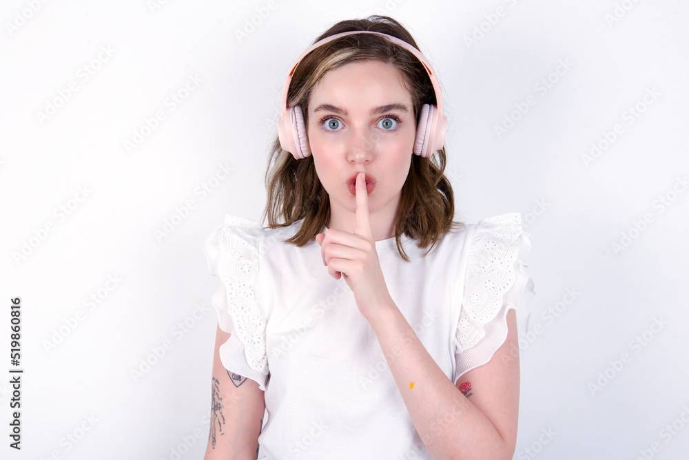 young caucasian woman wearing white T-shirt over white background making hush gesture with finger on her lips wearing wireless headphones. Be quiet. young caucasian woman wearing white T-shirt over white background making hush gesture with finger on her lips wearing wireless headphones. Be quiet.