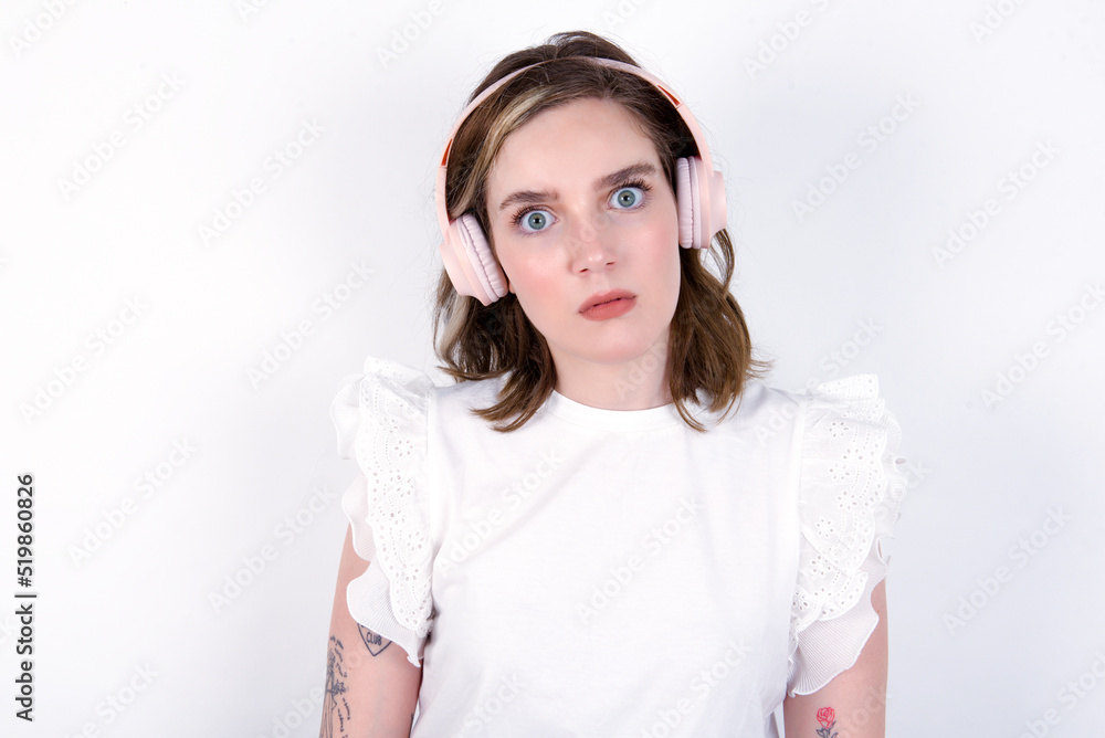 Serious displeased young caucasian woman wearing white T-shirt over white background looks puzzled at camera being angry wears stereo headphones listens music while walking at street Serious displeased young caucasian woman wearing white T-shirt over white background looks puzzled at camera being angry wears stereo headphones listens music while walking at street