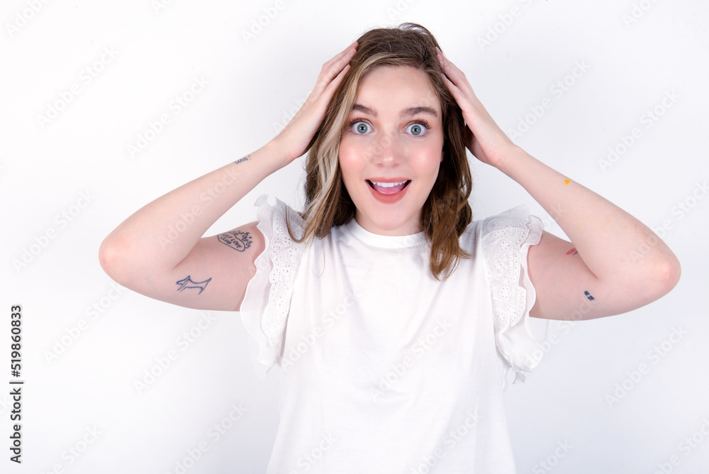 Cheerful overjoyed young caucasian woman wearing white T-shirt over white background reacts rising hands over head after receiving great news. Cheerful overjoyed young caucasian woman wearing white T-shirt over white background reacts rising hands over head after receiving great news.