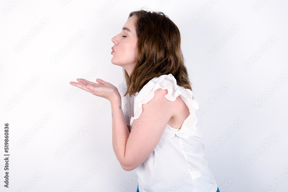 Profile side view, portrait of attractive young caucasian woman wearing white T-shirt over white background sending air kiss Profile side view, portrait of attractive young caucasian woman wearing white T-shirt over white background sending air kiss