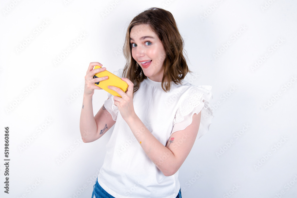 Nice addicted cheerful young caucasian woman wearing white T-shirt over white background using gadget playing network game