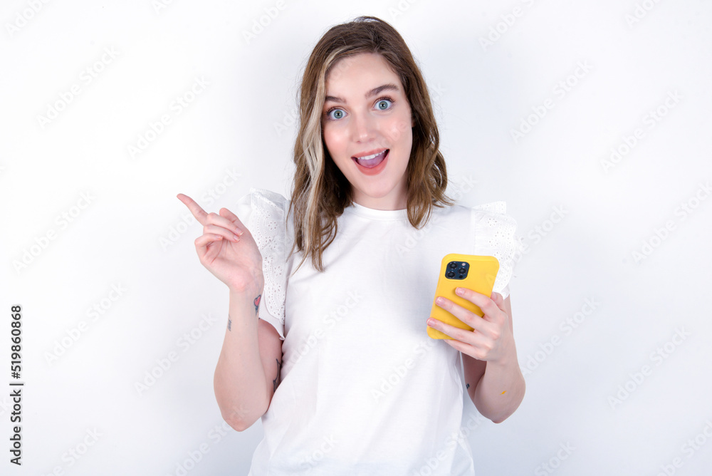 Astonished young caucasian woman wearing white T-shirt over white background holding her telephone and pointing with finger aside at empty copy space Astonished young caucasian woman wearing white T-shirt over white background holding her telephone and pointing with finger aside at empty copy space