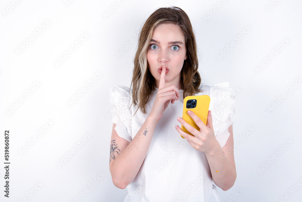 young caucasian woman wearing white T-shirt over white background holding modern gadget ask not tell secrets young caucasian woman wearing white T-shirt over white background holding modern gadget ask not tell secrets