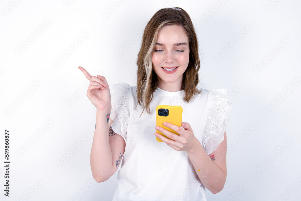 Smiling young caucasian woman wearing white T-shirt over white background pointing finger at blank space holding phone in one hand Smiling young caucasian woman wearing white T-shirt over white background pointing finger at blank space holding phone in one hand