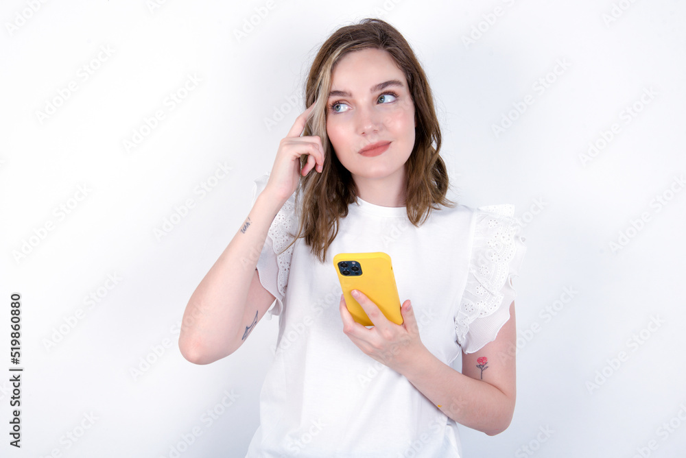 Pensive young caucasian woman wearing white T-shirt over white background keeps index finger on temple, holds modern smartphone and concentrated aside Pensive young caucasian woman wearing white T-shirt over white background keeps index finger on temple, holds modern smartphone and concentrated aside