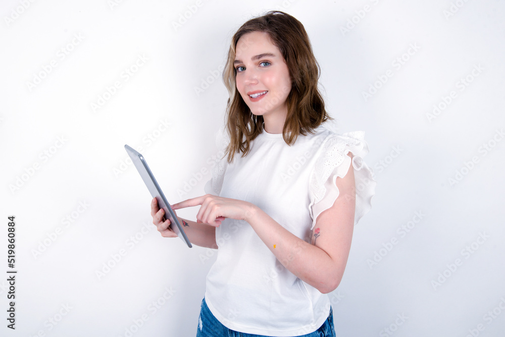 Photo of happy cheerful smart young caucasian woman wearing white T-shirt over white background hold tablet browsing internet Photo of happy cheerful smart young caucasian woman wearing white T-shirt over white background hold tablet browsing internet