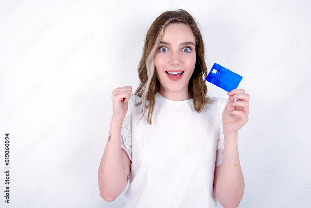 Photo of lucky impressed young caucasian woman wearing white T-shirt over white background arm fist holding credit card. Celebrated Photo of lucky impressed young caucasian woman wearing white T-shirt over white background arm fist holding credit card. Celebrated