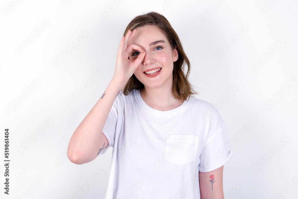 young caucasian woman wearing white T-shirt over white background with happy face smiling doing ok sign with hand on eye looking through finger. young caucasian woman wearing white T-shirt over white background with happy face smiling doing ok sign with hand on eye looking through finger.