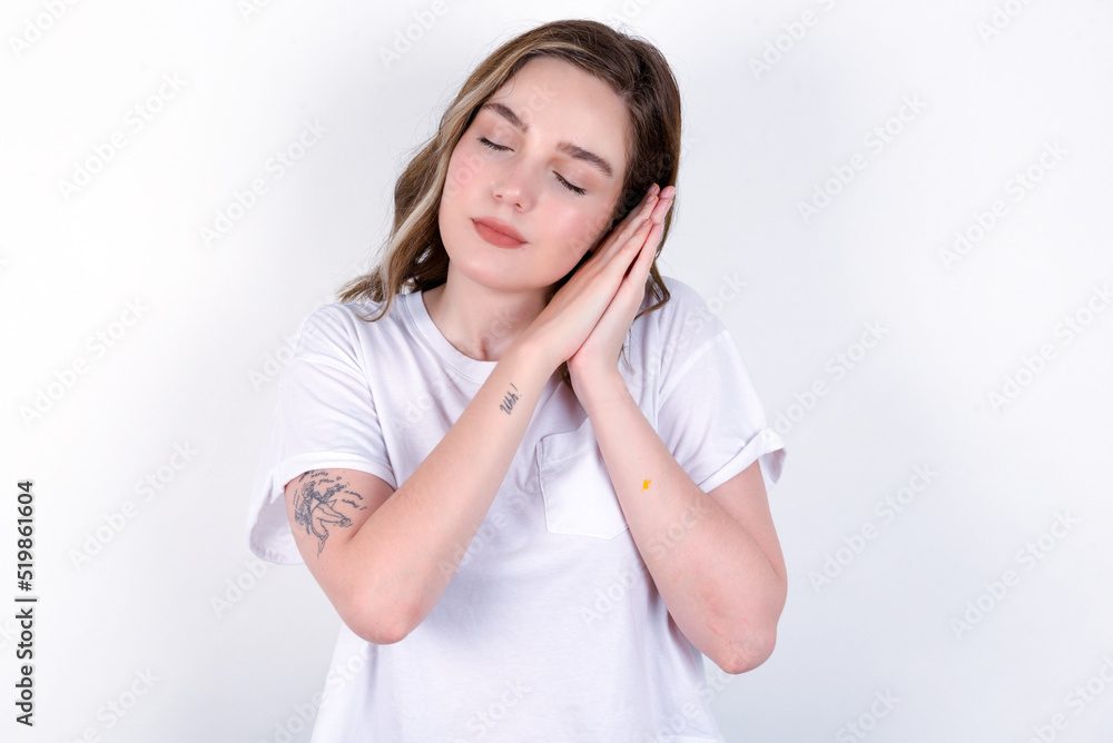 Relax and sleep time. Tired young caucasian woman wearing white T-shirt over white background with closed eyes leaning on palms making sleeping gesture. Relax and sleep time. Tired young caucasian woman wearing white T-shirt over white background with closed eyes leaning on palms making sleeping gesture.