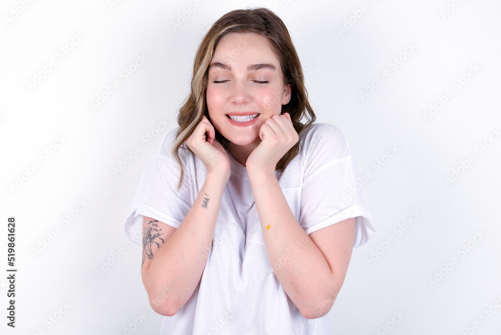 Portrait of young caucasian woman wearing white T-shirt over white background being overwhelmed, expressing excitement and happiness with closed eyes and hands near face. Portrait of young caucasian woman wearing white T-shirt over white background being overwhelmed, expressing excitement and happiness with closed eyes and hands near face.