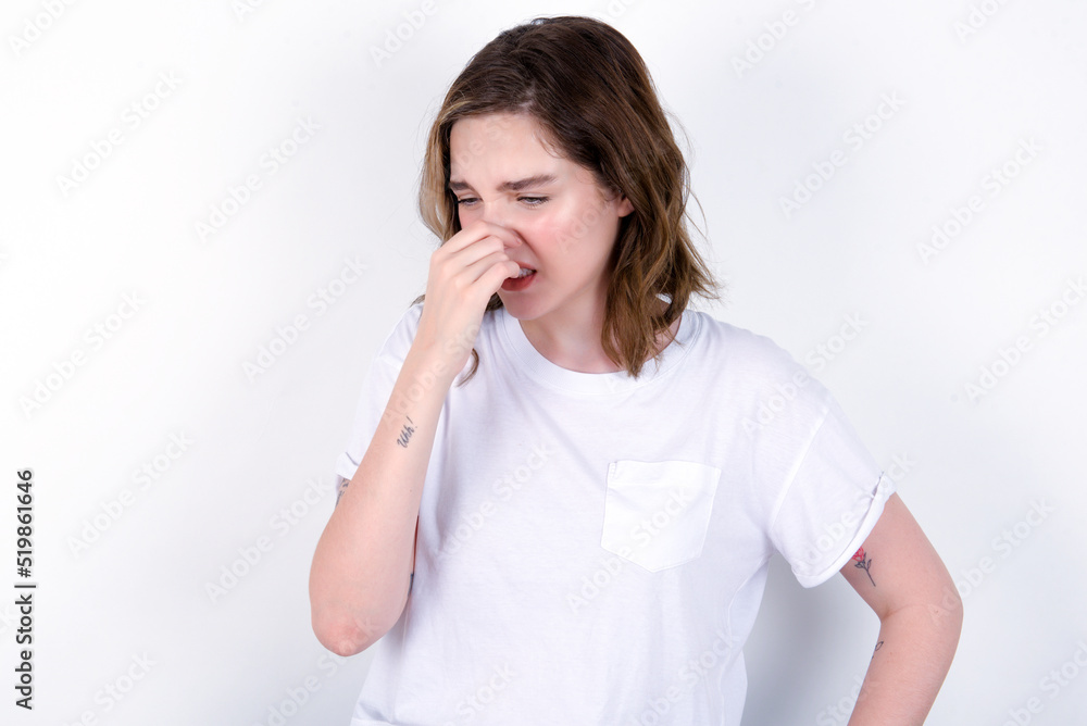 young caucasian woman wearing white T-shirt over white background , holding his nose because of a bad smell. young caucasian woman wearing white T-shirt over white background , holding his nose because of a bad smell.