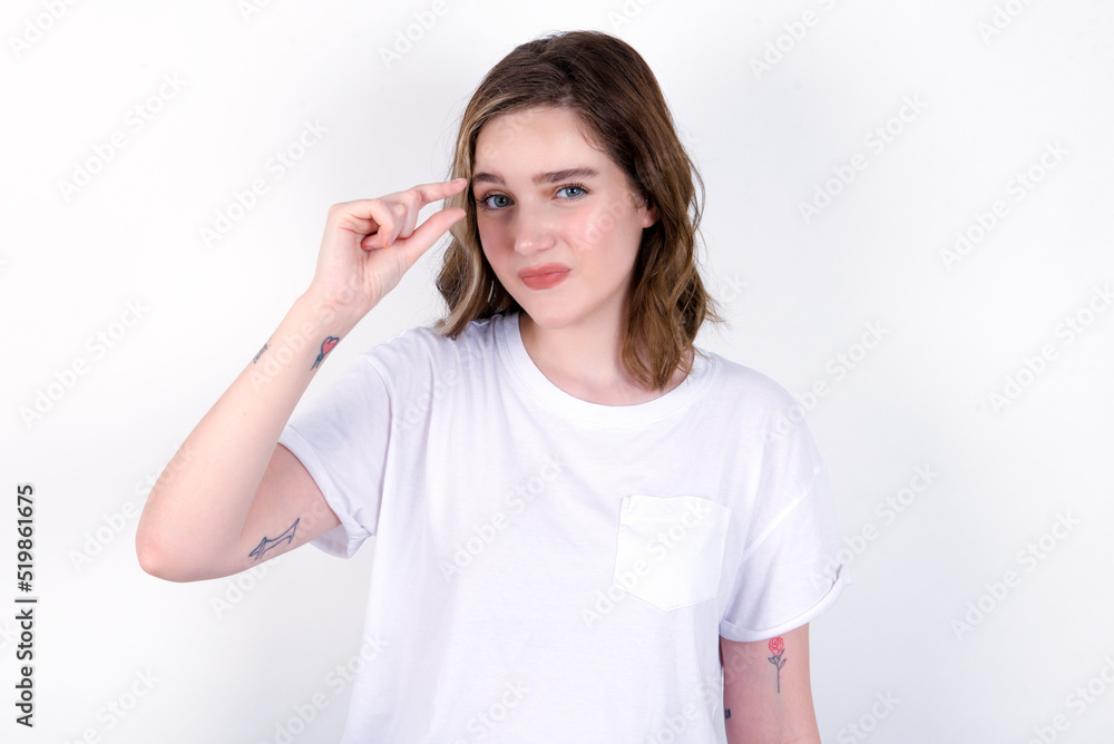 Upset young caucasian woman wearing white T-shirt over white background shapes little gesture with hand demonstrates something very tiny small size. Not very much Upset young caucasian woman wearing white T-shirt over white background shapes little gesture with hand demonstrates something very tiny small size. Not very much