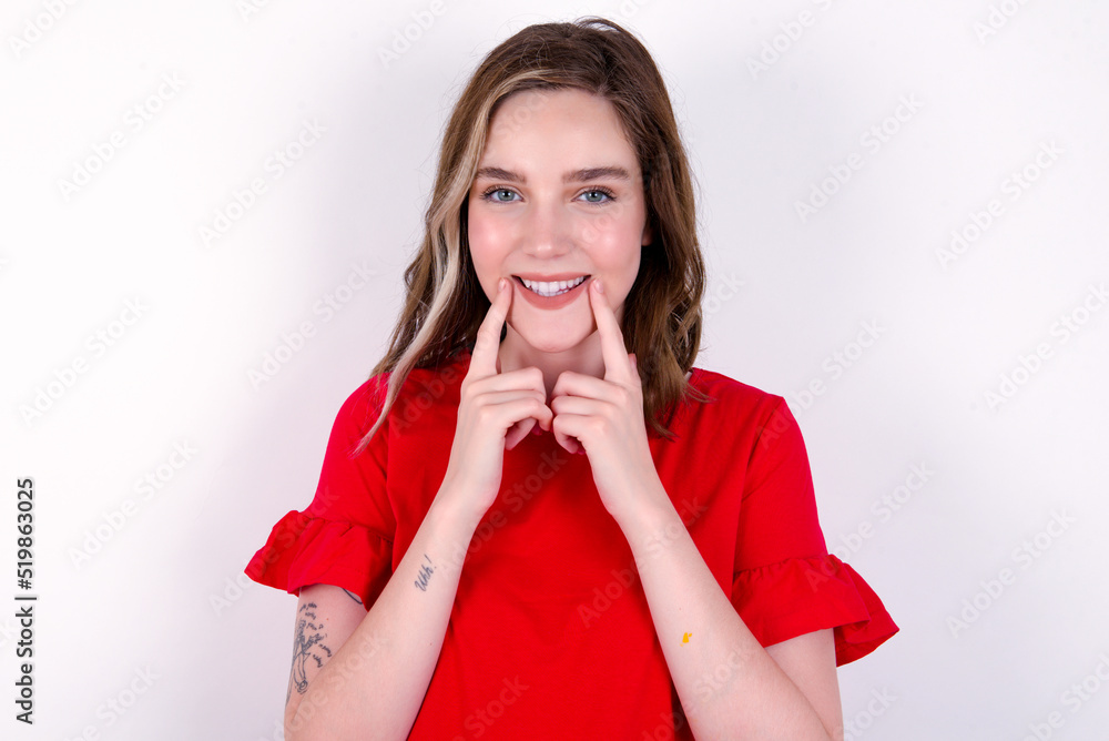 Happy young caucasian woman wearing red T-shirt over white background with toothy smile, keeps index fingers near mouth, fingers pointing and forcing cheerful smile Happy young caucasian woman wearing red T-shirt over white background with toothy smile, keeps index fingers near mouth, fingers pointing and forcing cheerful smile