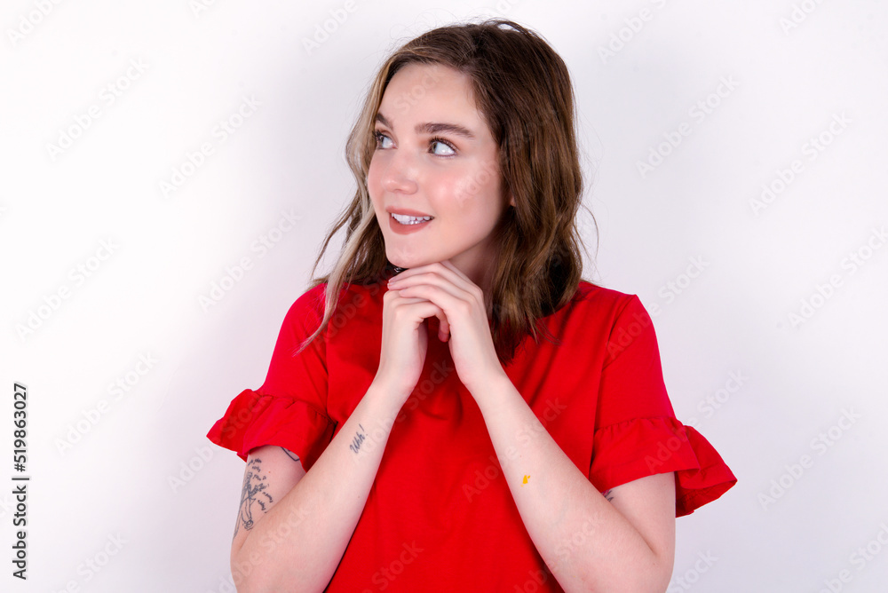 Happy young caucasian woman wearing red T-shirt over white background anticipates something awesome happen, looks happily aside, keeps hands together near face, has glad expression. Happy young caucasian woman wearing red T-shirt over white background anticipates something awesome happen, looks happily aside, keeps hands together near face, has glad expression.