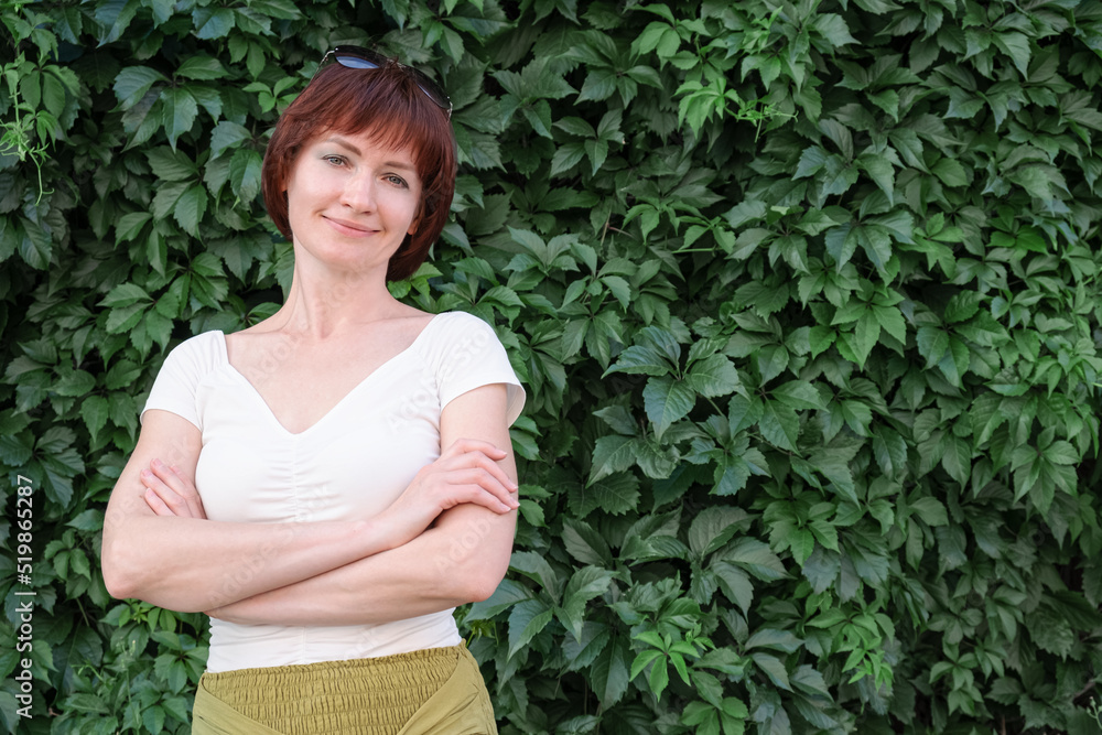 Woman with auburn hair stands with her arms crossed n front of a wall covered in leaves. Woman smiles and looks at the camera. Confident middle aged woman. Space for text Woman with auburn hair stands with her arms crossed n front of a wall covered in leaves. Woman smiles and looks at the camera. Confident middle aged woman. Space for text