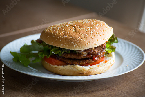 Homemade hamburger with double beef, tomato, green lettuce served on a white plate on a wooden table closeup view