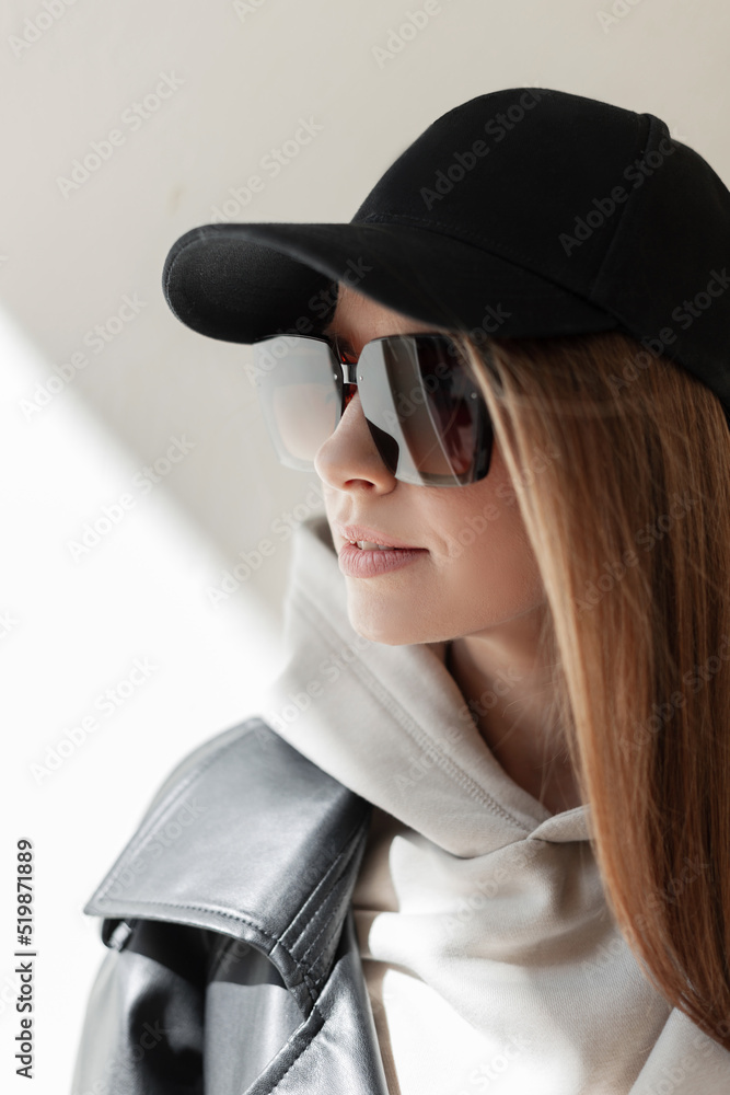 Urban female portrait of beautiful street fashion woman hipster with black cap and sunglasses in trendy leather coat and fashionable hoodie stands near a wall in sunlight on the street Urban female portrait of beautiful street fashion woman hipster with black cap and sunglasses in trendy leather coat and fashionable hoodie stands near a wall in sunlight on the street