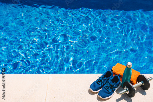 top view of sports objects in front of a swimming pool. orange towel, blue water bottle, weights. bottom of a swimming pool in a garden.