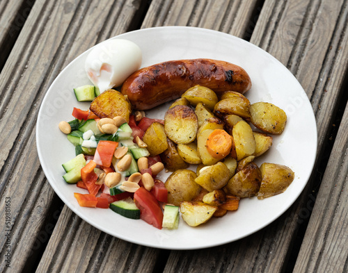 High angle view portion on a plate of delicious barbeque food on a wooden platform. Grilled sausage with rustic potato cubes, boiled egg and fresh salad.