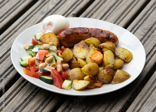 High angle view portion on a plate of delicious barbeque food on a wooden platform. Grilled sausage with rustic potato cubes, boiled egg and fresh salad.